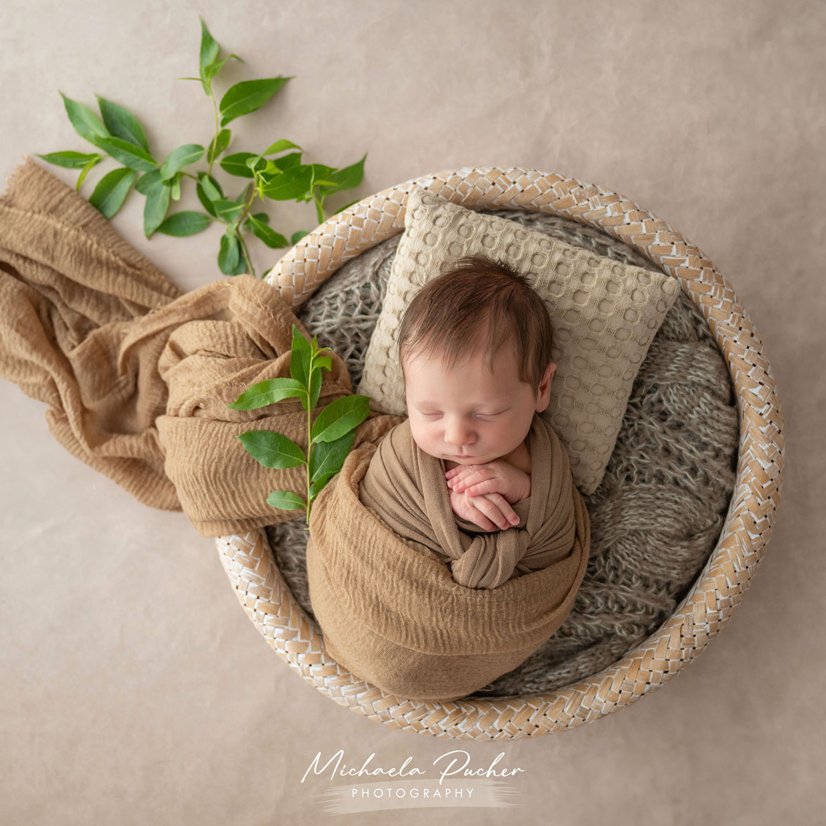 Schlafendes Neugeborenes, eingewickelt in braune Tücher, liegt in einem runden Korb auf weichen Decken; frische grüne Zweige dekorieren die Szene.